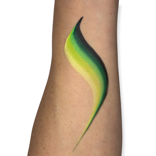 A close-up of a person's forearm with a face paint brush stroke in different shades of green on the skin, against a plain white background. The brush stroke looks like colors that can be used to create monster designs with face paint.