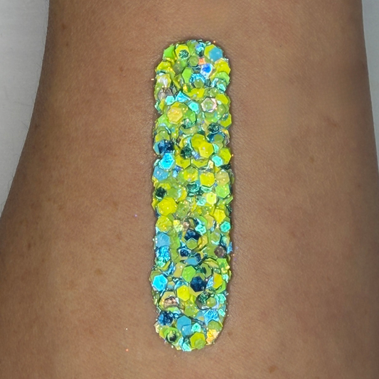 Close-up swatch of chunky cosmetic glitter cream in bright green, yellow, turquoise, and blue with iridescent shimmer, applied in a vertical stripe on skin. Inspired by the vibrant colors of Brazil