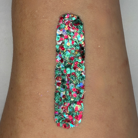 Close-up swatch of chunky cosmetic glitter cream applied in a vertical strip on skin. The glitter blend features vibrant emerald green, ruby red, holographic silver, and iridescent hexagonal glitter pieces in multiple sizes, creating a bold, festive sparkle with full coverage.