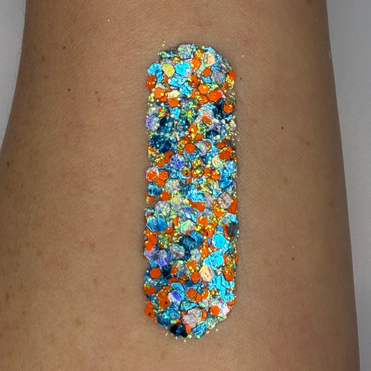 Bottom view of a 25g Studio Sparkle Glitter Cream jar showing the product label for Amplified. The clear round container reveals chunky blue, orange, silver, and iridescent cosmetic glitter around the edge, while the center label displays product name, ingredients, safety information, 12-month shelf life symbol, and SillyFarm manufacturing details on a white background.