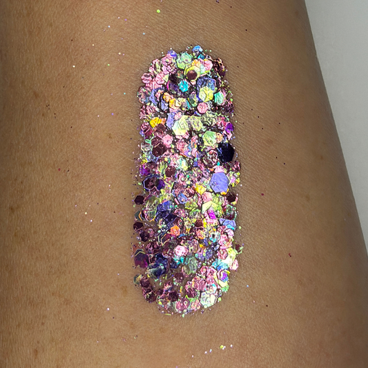 Close-up swatch of chunky cosmetic glitter applied to skin, featuring iridescent pink, lavender, and holographic hexagonal glitter pieces in multiple sizes. The glitter is densely packed in a vertical swatch, creating a magical, color-shifting sparkle effect with rainbow reflections and high-shine shimmer.