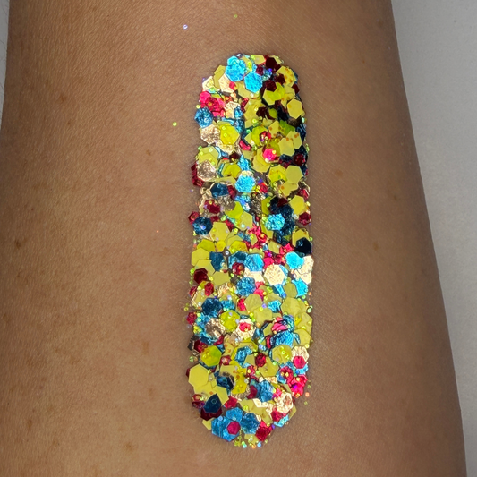 Close-up swatch of chunky cosmetic glitter applied to skin, featuring a bold mix of neon yellow, bright blue, hot pink, silver, and iridescent hexagonal glitter pieces in multiple sizes. The glitter is densely packed in a vertical swatch, creating a vibrant, high-shine sparkle effect inspired by soccer fan colors with reflective holographic highlights.