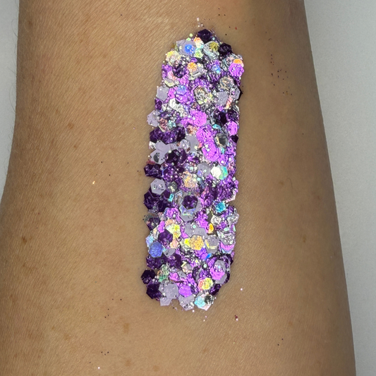 Close-up swatch of chunky cosmetic glitter applied to skin, featuring rich purple, lavender, and iridescent hexagonal glitter pieces in multiple sizes. The glitter is densely packed in a vertical swatch, creating a bold, sparkling effect with holographic rainbow reflections and high-shine shimmer.