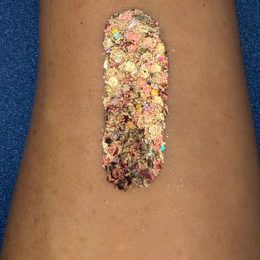 Close-up swatch of chunky cosmetic glitter applied to skin, featuring metallic gold, champagne, rose gold, and iridescent hexagonal glitter pieces in multiple sizes. The glitter is densely packed in a vertical swatch, creating a luxurious, sparkling effect with holographic rainbow reflections and warm metallic shine.