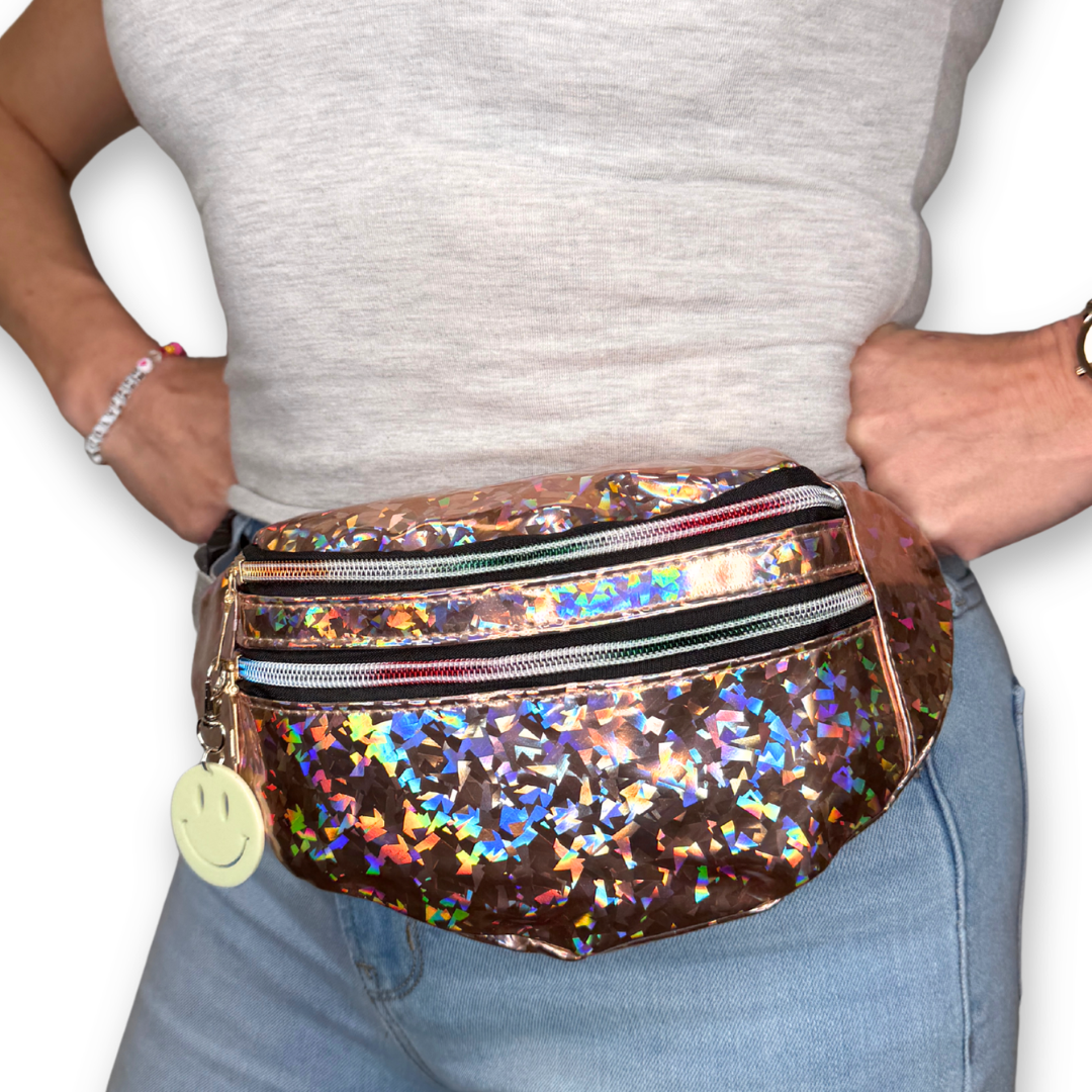 A person wearing a shiny champagne holographic fanny pack with a smiley face charm, styled with a gray tank top and light blue jeans against a white background.