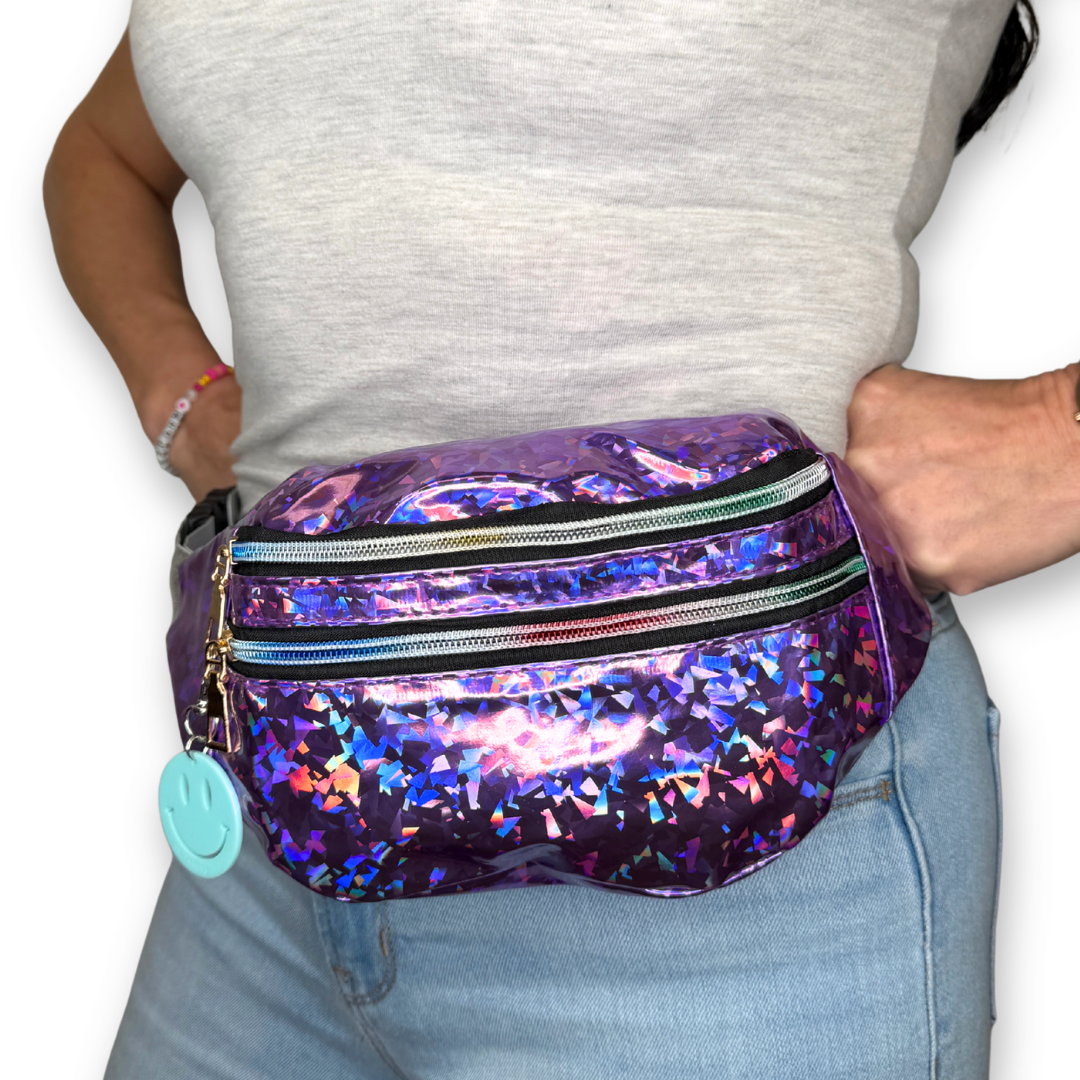 A person wearing a shiny holographic purple fanny pack with multiple zippered compartments and a small light blue smiley face charm, shown against a plain white background.