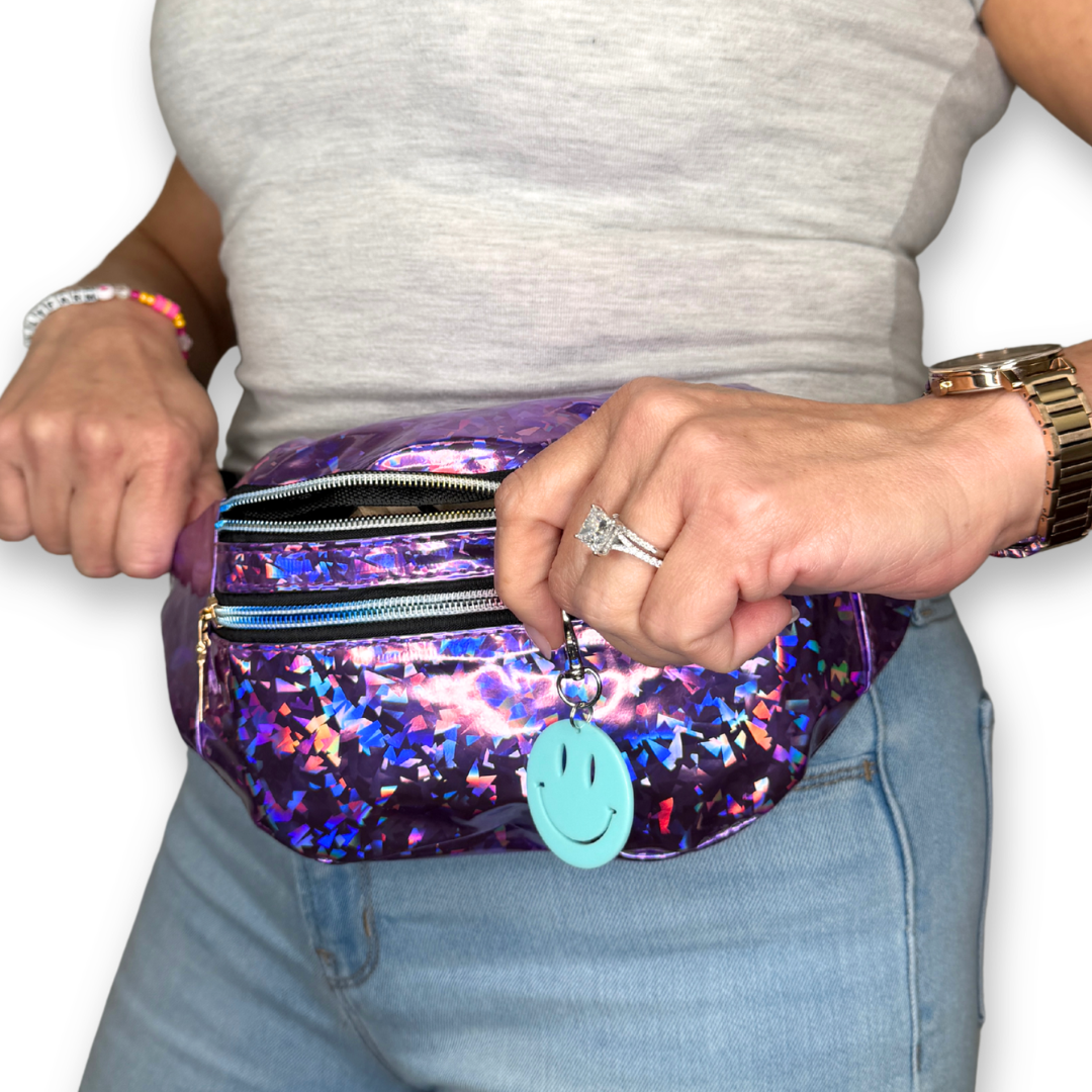 A person wearing a shiny holographic purple fanny pack with star patterns, a light blue smiley face charm, and gold zippers, paired with a gray tank top and blue jeans.