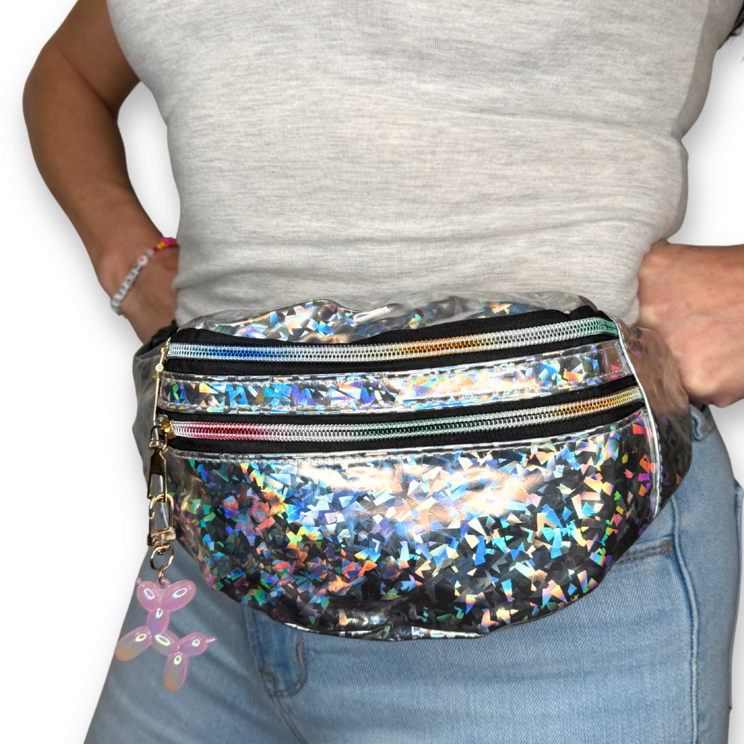 A person wearing a shiny holographic fanny pack with multiple zippered compartments and a small pink balloon-shaped charm, shown from the chest down against a plain white background.