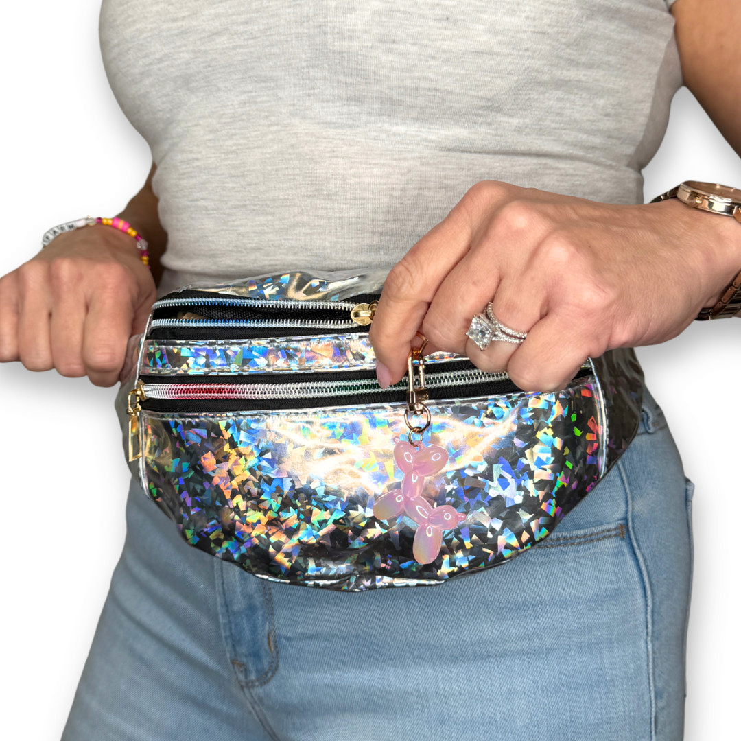 A person wearing a shiny holographic fanny pack with multiple zip compartments and a pink bear charm, shown from the chest down against a white background.