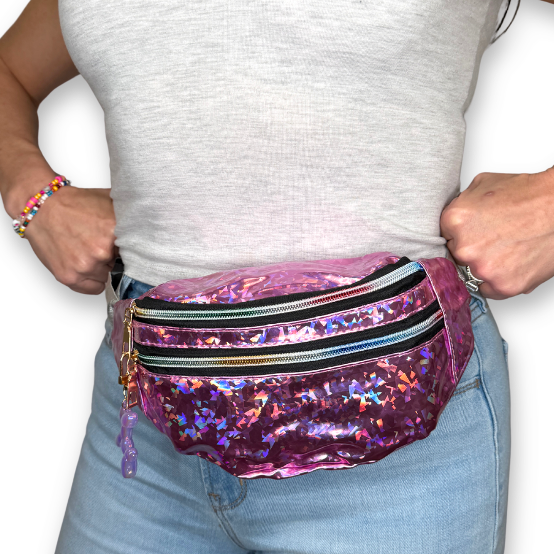 A person wearing a shiny holographic light pink fanny pack or belt bag over light blue jeans and a light gray t-shirt, with both hands pulling the bag out and adjusting it.