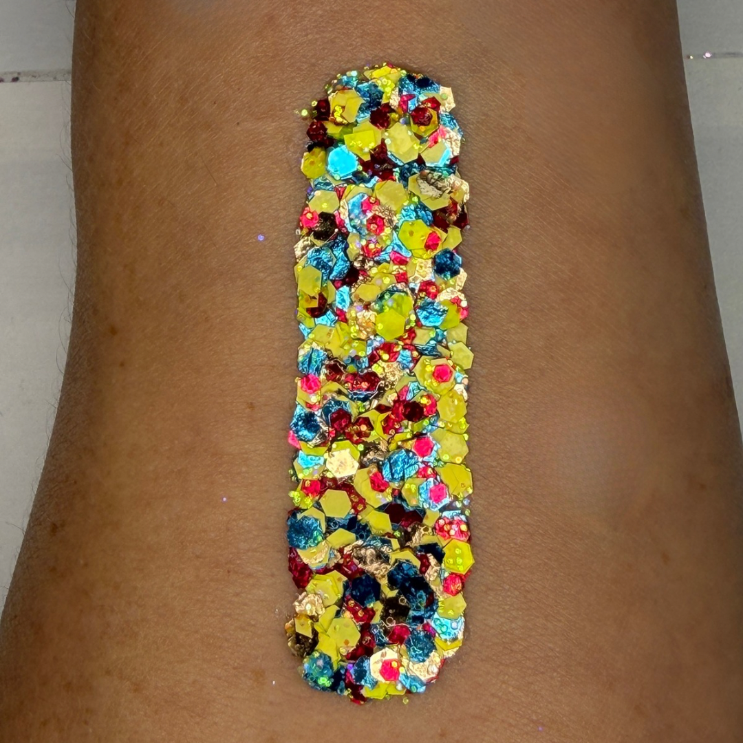 Close-up swatch of chunky cosmetic glitter cream applied in a vertical strip on skin. The glitter blend features bright yellow, turquoise blue, ruby red, holographic silver, and iridescent hexagonal glitter pieces in multiple sizes, creating a vibrant, high-shine sparkle with bold, full coverage.