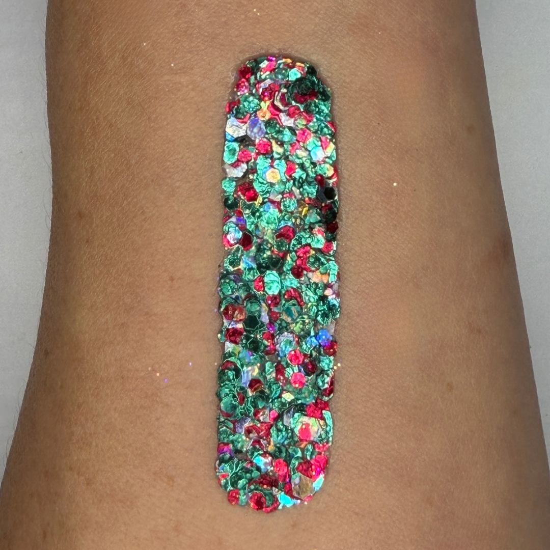 Close-up swatch of chunky cosmetic glitter cream applied in a vertical strip on skin. The glitter blend features vibrant emerald green, ruby red, holographic silver, and iridescent hexagonal glitter pieces in multiple sizes, creating a bold, festive sparkle with full coverage.