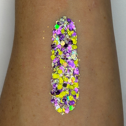 Close-up swatch of chunky cosmetic glitter applied to skin, featuring neon yellow, purple, silver, and iridescent hexagonal glitter pieces in multiple sizes. The glitter is densely packed in a vertical swatch, creating a bold, cosmic-inspired sparkle effect with holographic rainbow reflections and high-shine shimmer.