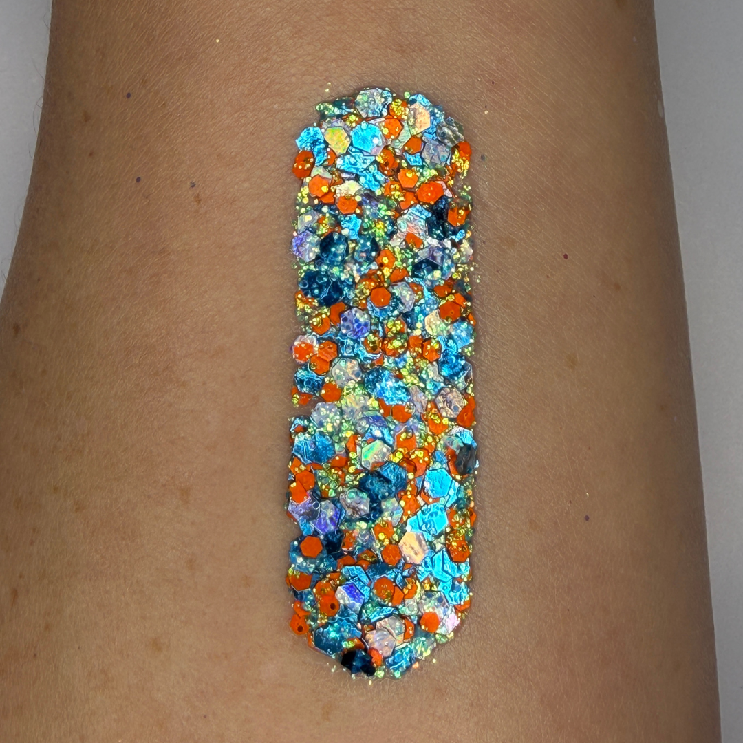 Bottom view of a 25g Studio Sparkle Glitter Cream jar showing the product label for Amplified. The clear round container reveals chunky blue, orange, silver, and iridescent cosmetic glitter around the edge, while the center label displays product name, ingredients, safety information, 12-month shelf life symbol, and SillyFarm manufacturing details on a white background.