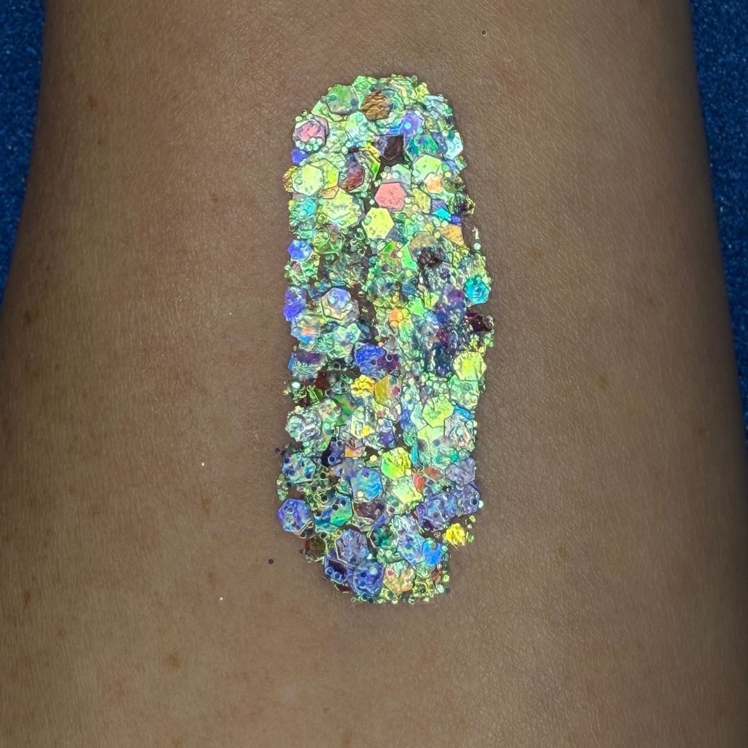 Close-up swatch of chunky cosmetic glitter applied to skin, featuring iridescent silver, aqua, blues, and holographic hexagonal glitter pieces in multiple sizes. The glitter is densely packed in a vertical swatch, creating a luminous, color-shifting sparkle effect with reflective rainbow highlights.