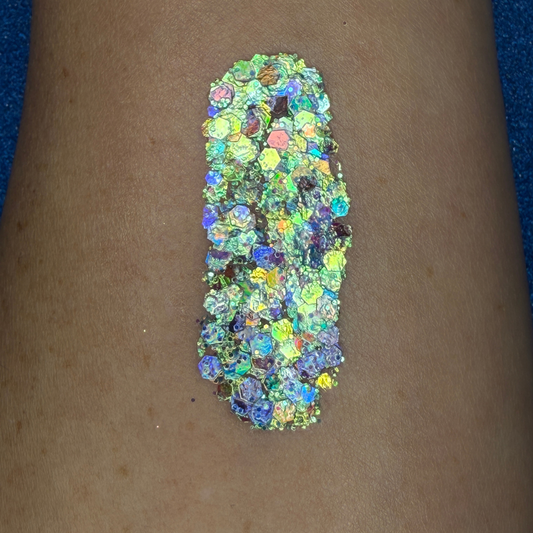 Close-up swatch of chunky cosmetic glitter applied to skin, featuring iridescent silver, aqua, blues, and holographic hexagonal glitter pieces in multiple sizes. The glitter is densely packed in a vertical swatch, creating a luminous, color-shifting sparkle effect with reflective rainbow highlights.