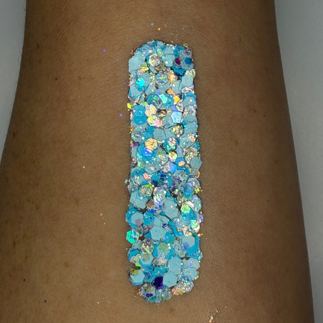 Close-up swatch of chunky cosmetic glitter applied to skin, featuring icy blue, aqua, white, and iridescent hexagonal glitter pieces in multiple sizes. The glitter is densely packed in a vertical swatch, creating a frosty, ocean-inspired sparkle effect with holographic reflections and shimmering rainbow highlights.