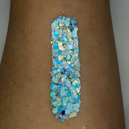 Close-up swatch of chunky cosmetic glitter applied to skin, featuring icy blue, aqua, white, and iridescent hexagonal glitter pieces in multiple sizes. The glitter is densely packed in a vertical swatch, creating a frosty, ocean-inspired sparkle effect with holographic reflections and shimmering rainbow highlights.