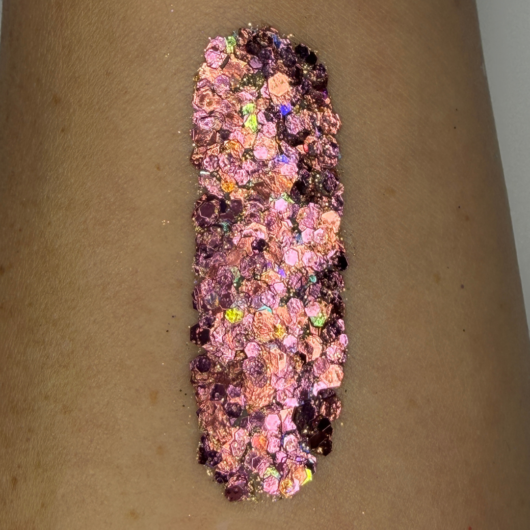 Close-up swatch of chunky cosmetic glitter applied to skin, featuring rose gold, blush pink, and iridescent hexagonal glitter pieces in multiple sizes. The glitter is densely packed in a vertical swatch, creating a luxurious sparkling effect with warm metallic shine and holographic rainbow reflections.