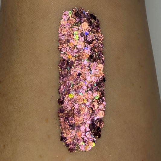 Close-up swatch of chunky cosmetic glitter applied to skin, featuring rose gold, blush pink, and iridescent hexagonal glitter pieces in multiple sizes. The glitter is densely packed in a vertical swatch, creating a luxurious sparkling effect with warm metallic shine and holographic rainbow reflections.