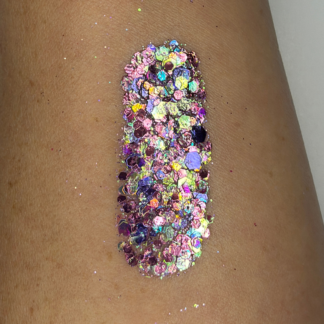 Close-up swatch of chunky cosmetic glitter applied to skin, featuring iridescent pink, lavender, and holographic hexagonal glitter pieces in multiple sizes. The glitter is densely packed in a vertical swatch, creating a magical, color-shifting sparkle effect with rainbow reflections and high-shine shimmer.