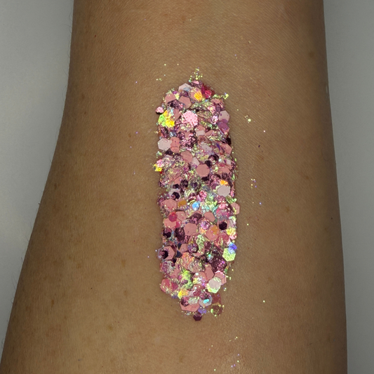 Close-up swatch of chunky cosmetic glitter applied to skin, featuring soft pink, rose pink, and iridescent hexagonal glitter pieces in multiple sizes. The glitter is densely packed in a vertical swatch, creating a sparkling pastel effect with holographic rainbow reflections and high-shine shimmer.
