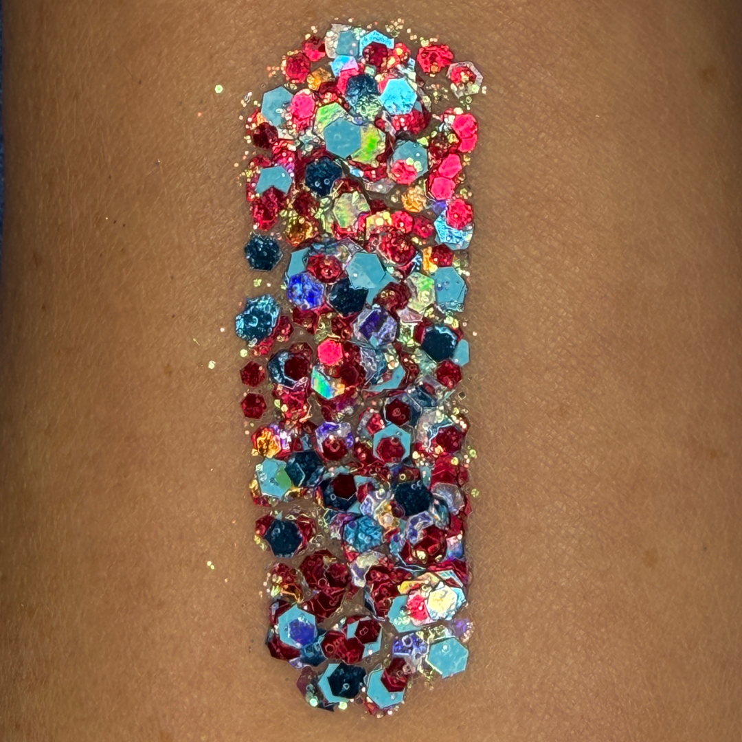 Close-up swatch of chunky cosmetic glitter applied to skin, featuring bright red, blue, and iridescent hexagonal glitter pieces in multiple sizes. The glitter is densely packed in a vertical swatch, creating a bold, patriotic-inspired sparkle effect with holographic rainbow reflections and high-shine shimmer.