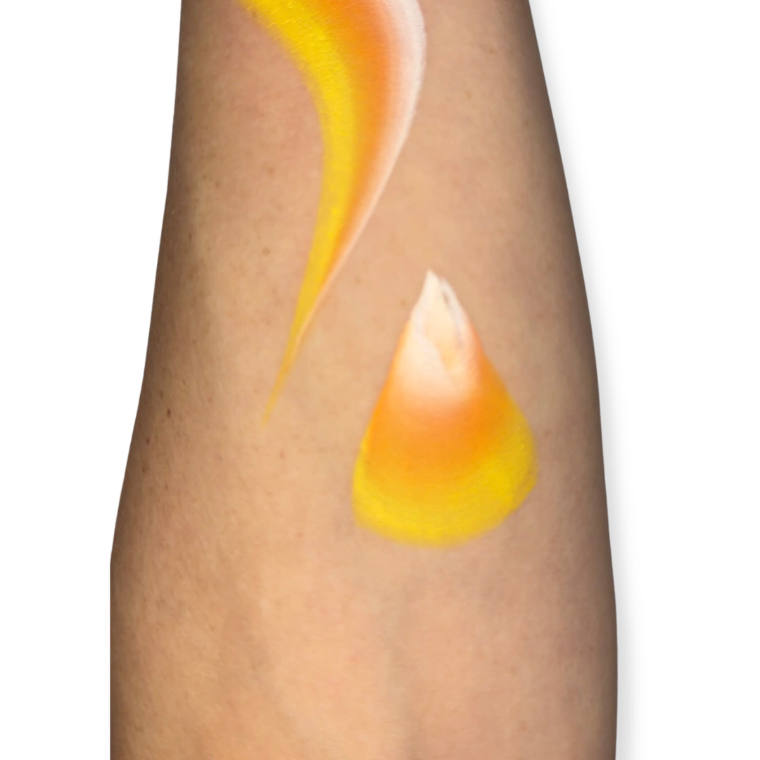 A close-up of a person's forearm with a face paint brush stroke in yellow, orange, and white on the skin, against a plain white background. The brush stroke looks like candy corn. 