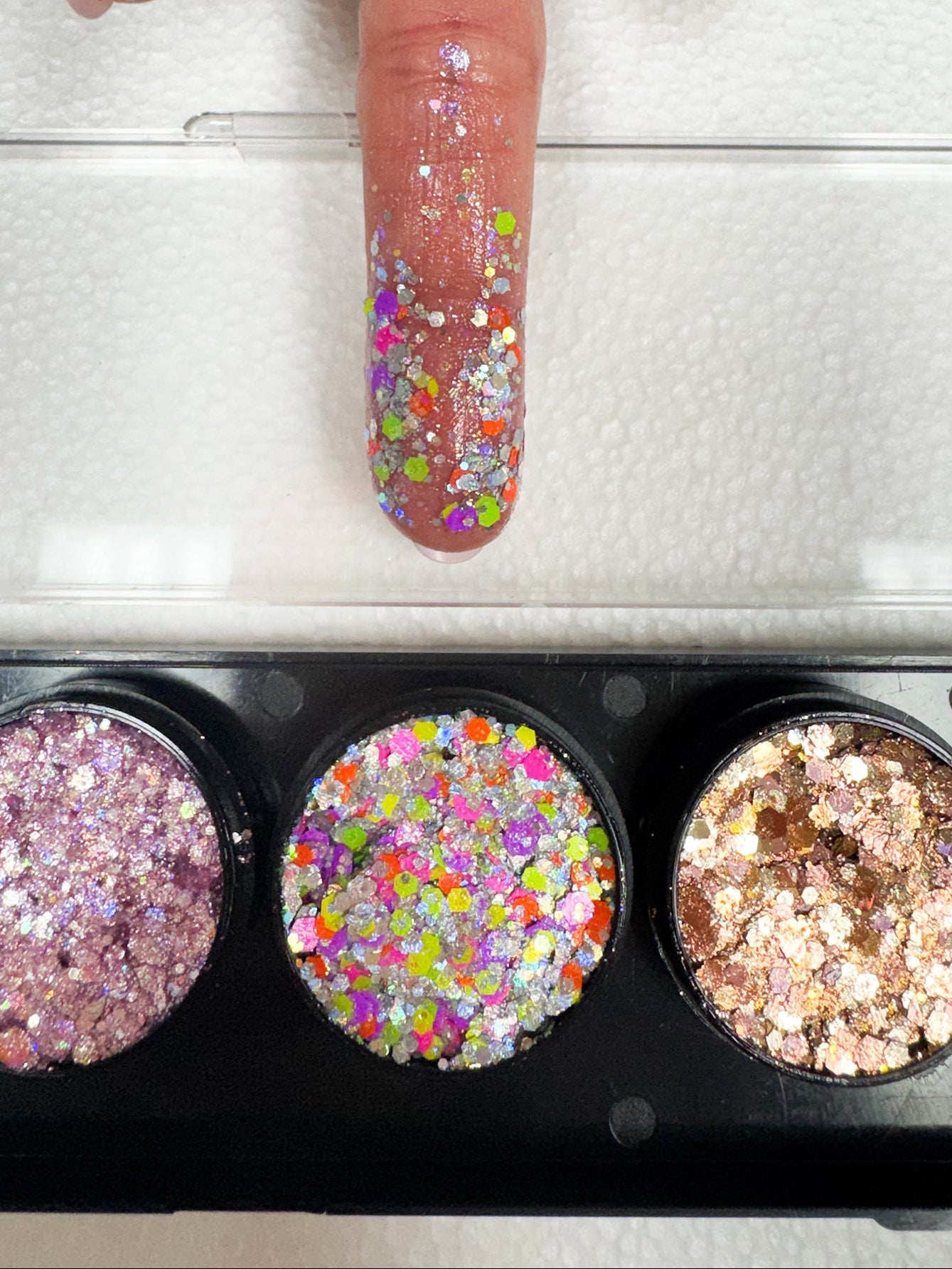 Finger with multicolored glittery nail polish above a glittery nail polish tray.