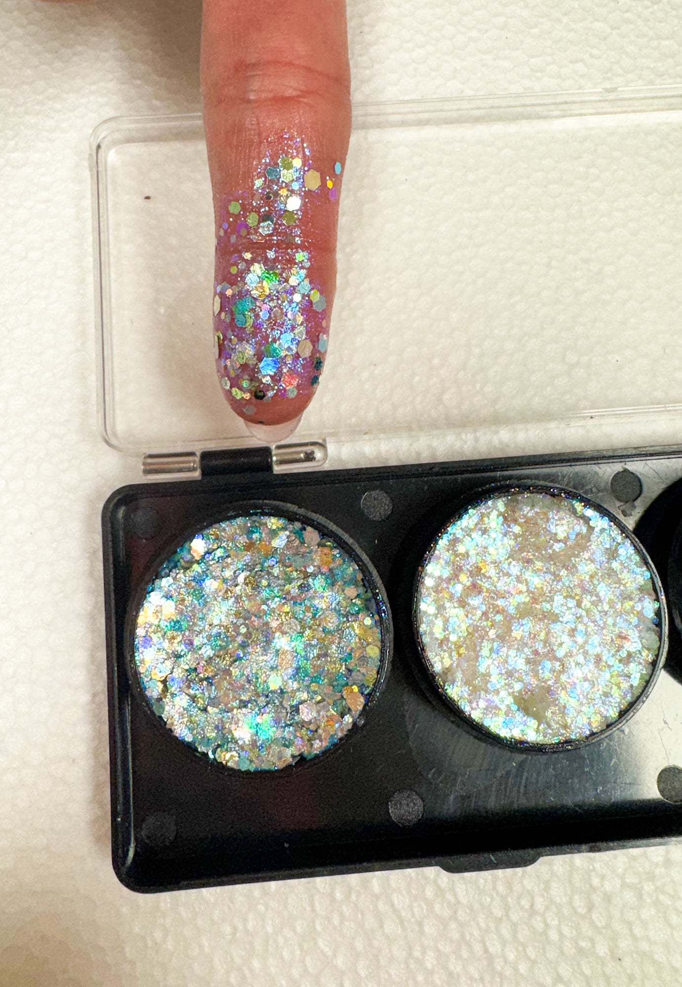 Eyeshadow palette with glittery shades and a finger with glittery nail polish on a light background