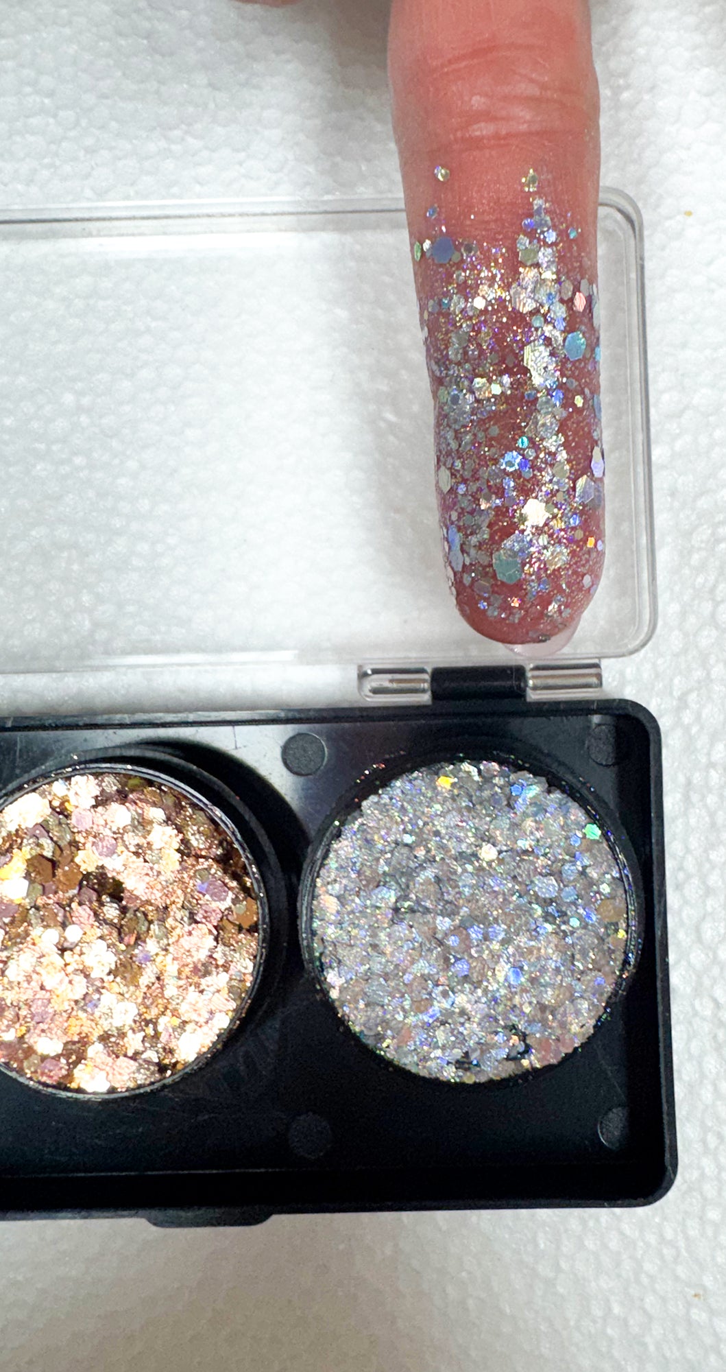 Two glittery eyeshadows in a compact with a finger showing glittery nail polish.