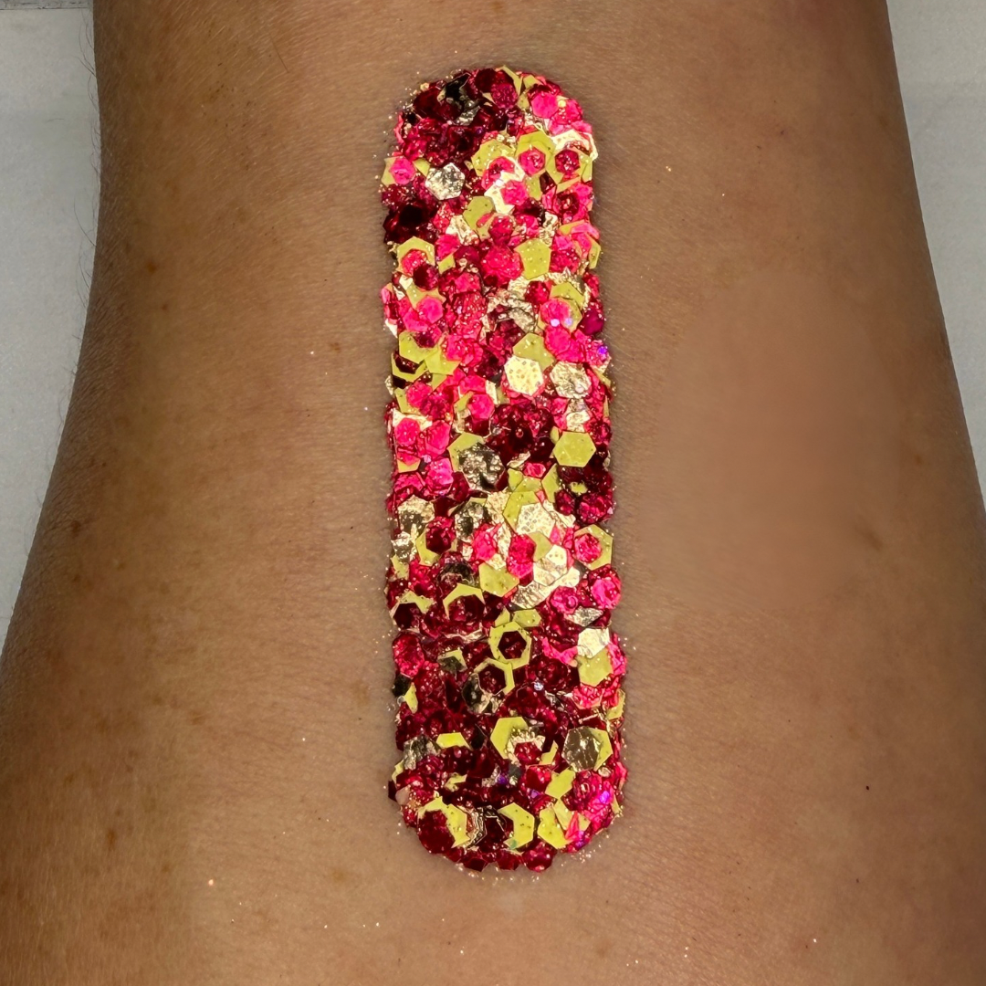 Close-up swatch of chunky cosmetic glitter cream applied in a vertical strip on skin. The glitter blend features hot pink, deep ruby red, metallic gold, and iridescent hexagonal glitter pieces in multiple sizes, creating a bold, dazzling sparkle with rich, full coverage.