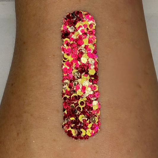 Close-up swatch of chunky cosmetic glitter cream applied in a vertical strip on skin. The glitter blend features hot pink, deep ruby red, metallic gold, and iridescent hexagonal glitter pieces in multiple sizes, creating a bold, dazzling sparkle with rich, full coverage.